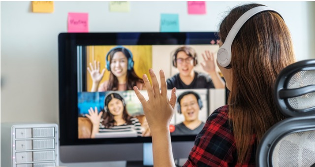 Woman wearing headphones participating in an online accent modification class, waving to four classmates on a video call.