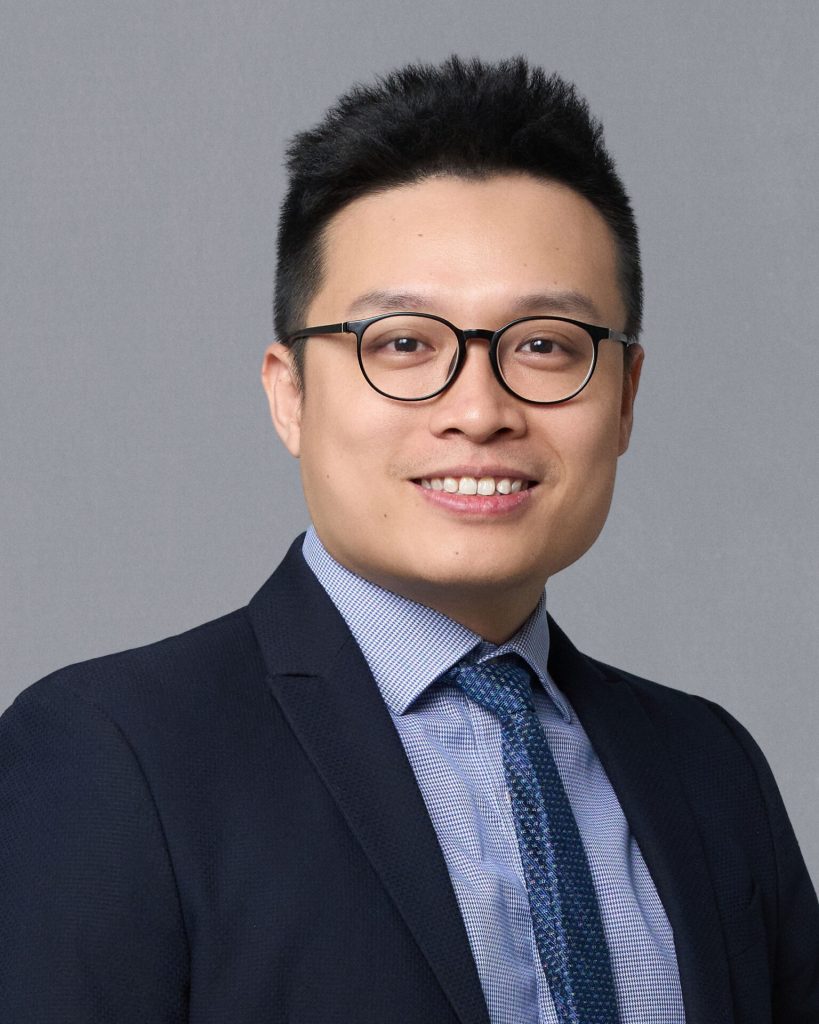 Portrait of a professional man in a dark suit and tie, smiling confidently. He is wearing glasses and is set against a neutral gray background.