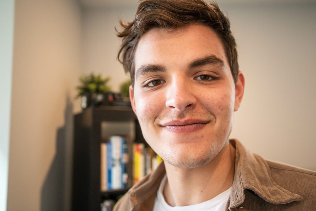 Close-up portrait of a young millennial businessman with a light smile, wearing a brown shirt and white t-shirt. He is standing indoors with bookshelves in the background and a plant visible. The image conveys a friendly and approachable demeanor.